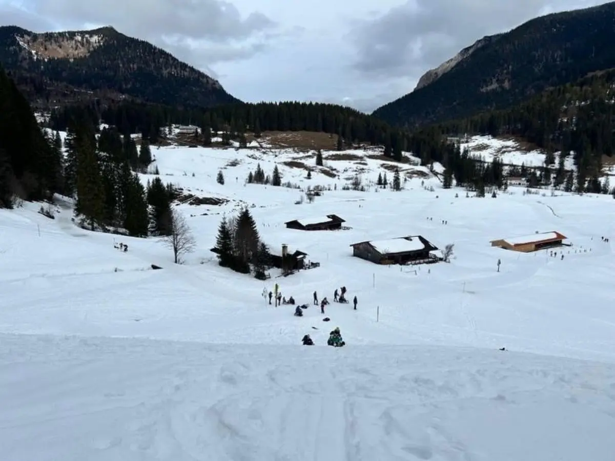 Verschneites Tal mit verstreuten Holzhütten; unten bewegen sich mehrere Menschen beim Aufstieg, umgeben von Wald und Bergkulisse unter grauem Himmel. | © DAV Markt Schwaben