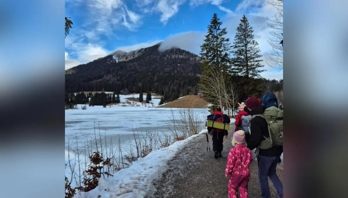 Kinder und Erwachsene gehen am verschneiten Uferweg entlang eines gefrorenen Sees; im Hintergrund erheben sich Berge und Wald unter blauem Himmel. | © DAV Markt Schwaben