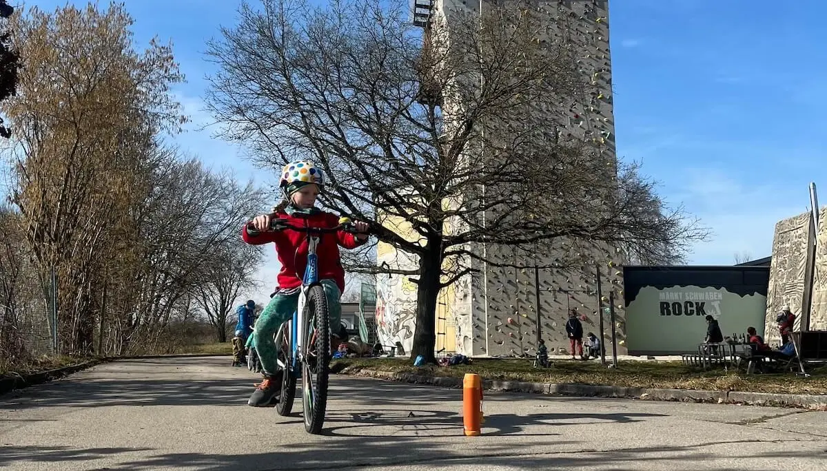 Ein Kind fährt im Vordergrund mit Helm und roter Jacke auf einem Radweg, während im Hintergrund weitere Kinder auf Fahrrädern üben. | © DAV Markt Schwaben/Miron Thoms