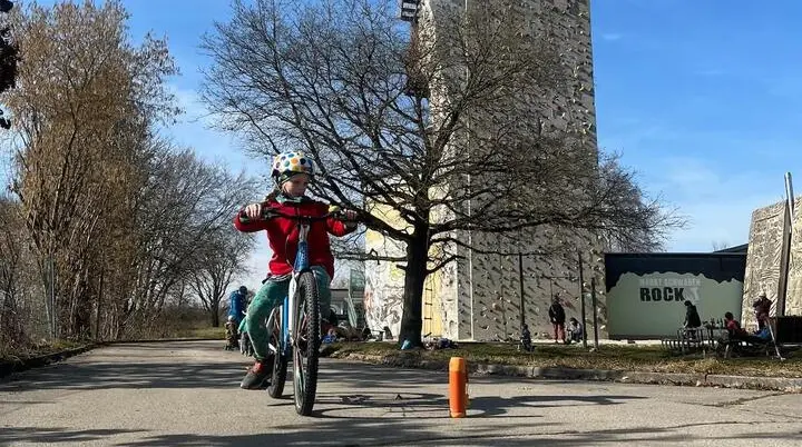 Ein Kind fährt im Vordergrund mit Helm und roter Jacke auf einem Radweg, während im Hintergrund weitere Kinder auf Fahrrädern üben. | © DAV Markt Schwaben / Miron Thoms