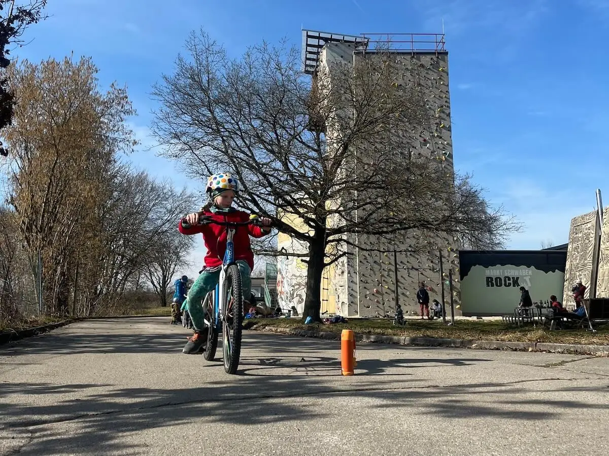 Ein Kind fährt im Vordergrund mit Helm und roter Jacke auf einem Radweg, während im Hintergrund weitere Kinder auf Fahrrädern üben. | © DAV Markt Schwaben/Miron Thoms