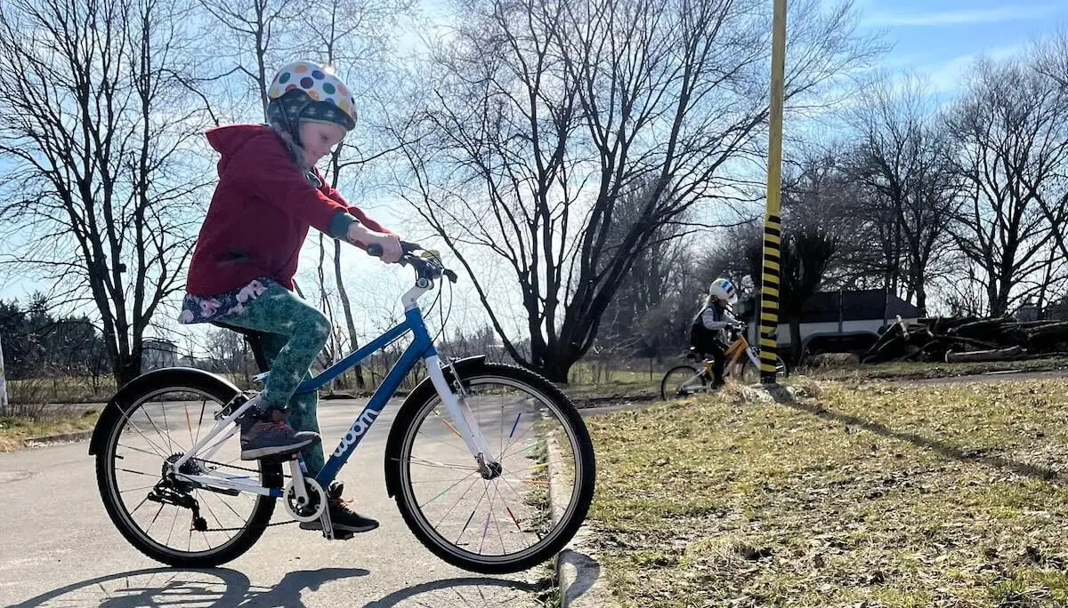Ein Kind fährt im Vordergrund mit Helm und roter Jacke auf einem blauen Fahrrad, während ein weiteres Kind im Hintergrund an einem gelb‑schwarzen Pfosten vorbeiradelt. | © DAV Markt Schwaben/Miron Thoms
