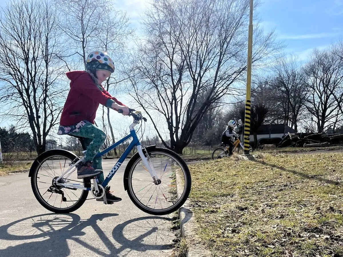 Ein Kind fährt im Vordergrund mit Helm und roter Jacke auf einem blauen Fahrrad, während ein weiteres Kind im Hintergrund an einem gelb‑schwarzen Pfosten vorbeiradelt. | © DAV Markt Schwaben/Miron Thoms