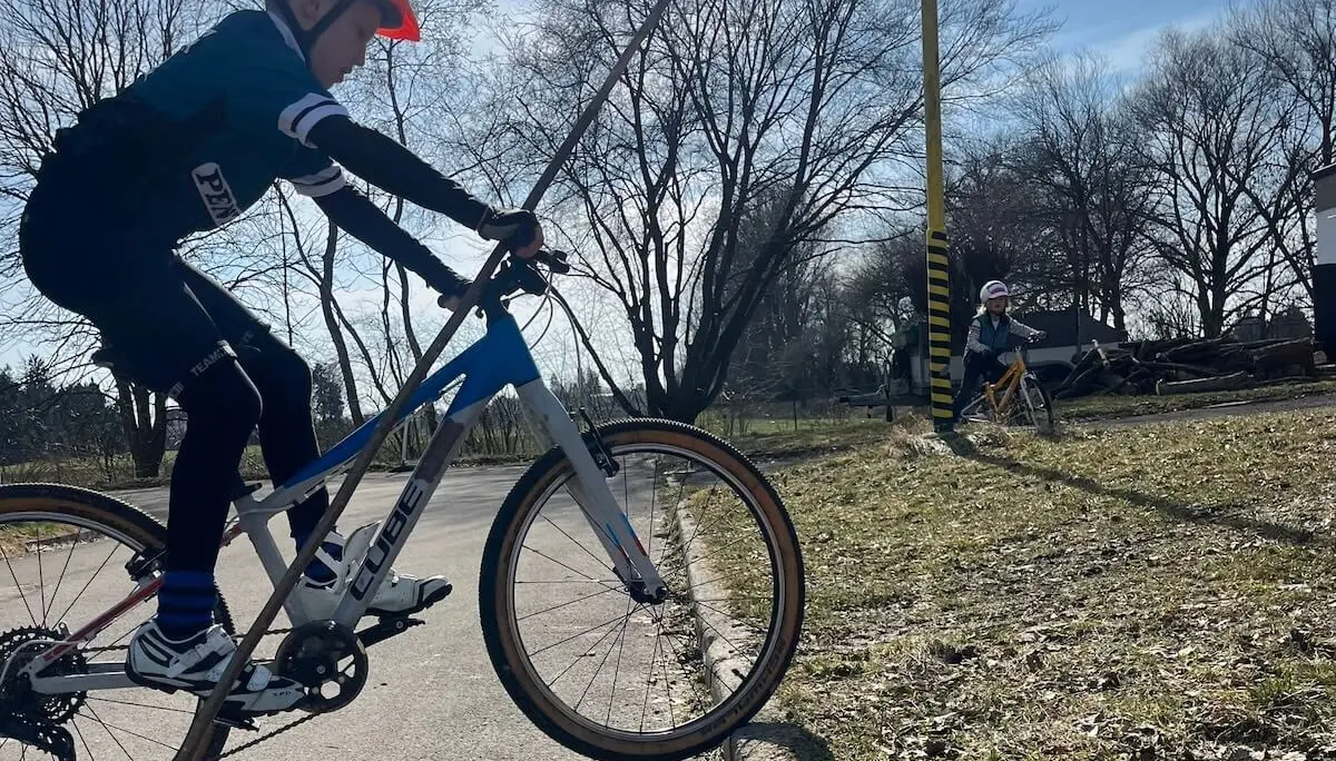 Ein Kind fährt im Vordergrund auf einem weiß‑blauen Mountainbike, während ein weiteres Kind im Hintergrund auf einem gelben Rad neben einem markierten Pfosten unterwegs ist. | © DAV Markt Schwaben/Miron Thoms