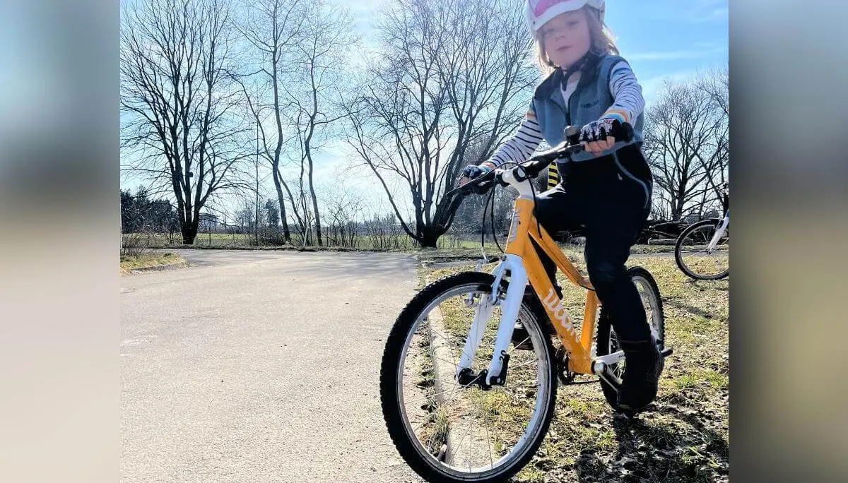 Ein Kind steht mit Helm und Handschuhen auf einem gelb‑weißen Fahrrad am Wegesrand, während im Hintergrund ein weiteres Rad neben kahlen Bäumen erkennbar ist. | © DAV Markt Schwaben/Miron Thoms