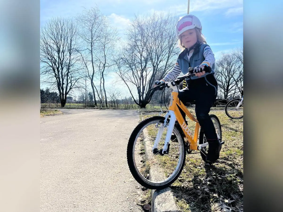 Ein Kind steht mit Helm und Handschuhen auf einem gelb‑weißen Fahrrad am Wegesrand, während im Hintergrund ein weiteres Rad neben kahlen Bäumen erkennbar ist. | © DAV Markt Schwaben/Miron Thoms