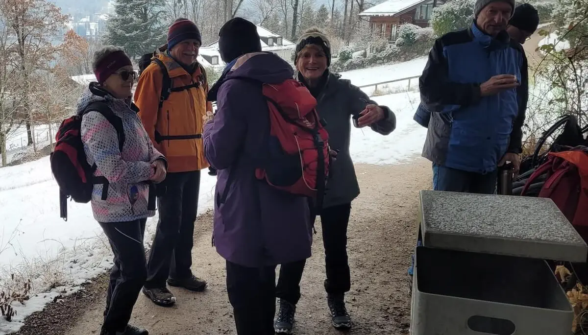 Sechs Wandernde stehen auf einem verschneiten Weg; vorne ein Abfallbehälter, dahinter Häuser, Bäume und winterliche Hänge.  | © DAV Markt Schwaben/Hubert Inhofer