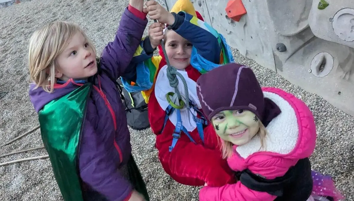 Drei verkleidete Kinder halten gemeinsam ein Kletterseil, lachen in die Kamera und stehen vor einer Kletterwand mit bunten Griffen. | © DAV Markt Schwaben