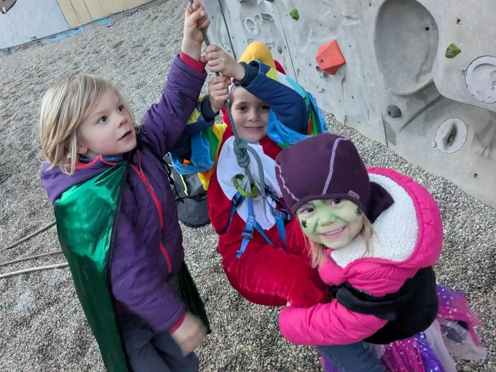 Drei verkleidete Kinder halten gemeinsam ein Kletterseil, lachen in die Kamera und stehen vor einer Kletterwand mit bunten Griffen. | © DAV Markt Schwaben