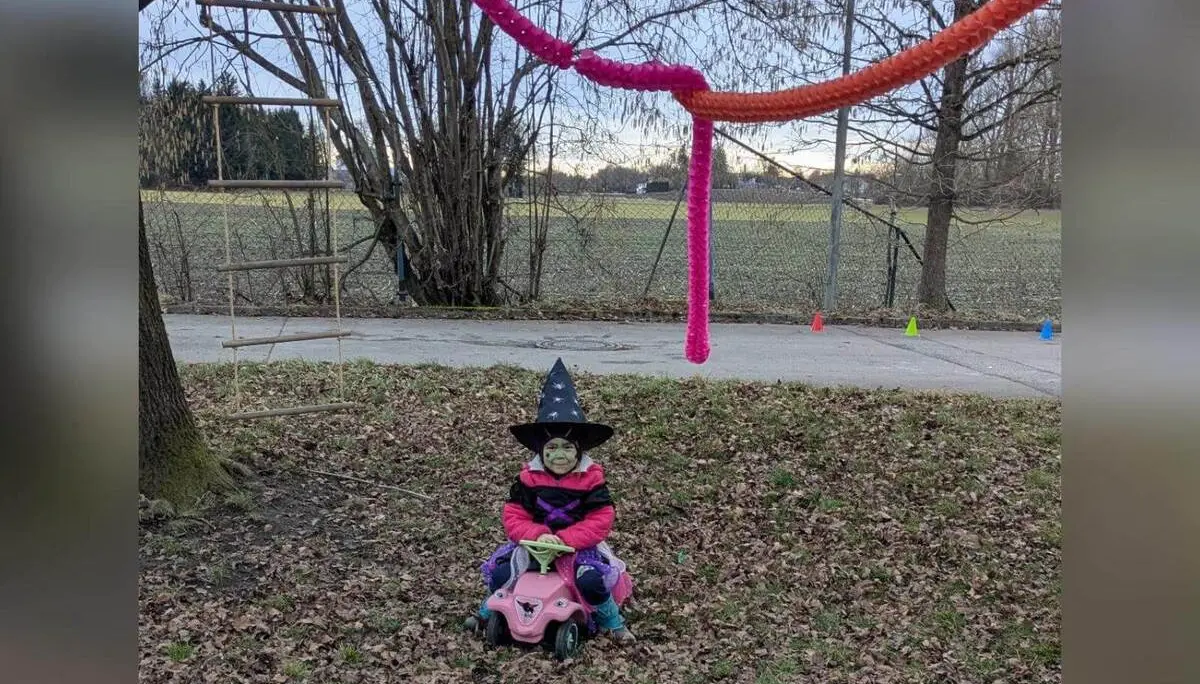 Ein verkleidetes kleines Kind mit Hexenhut sitzt auf einem Rutschfahrzeug unter bunten Girlanden an einem Baum auf einer Wiese. | © DAV Markt Schwaben