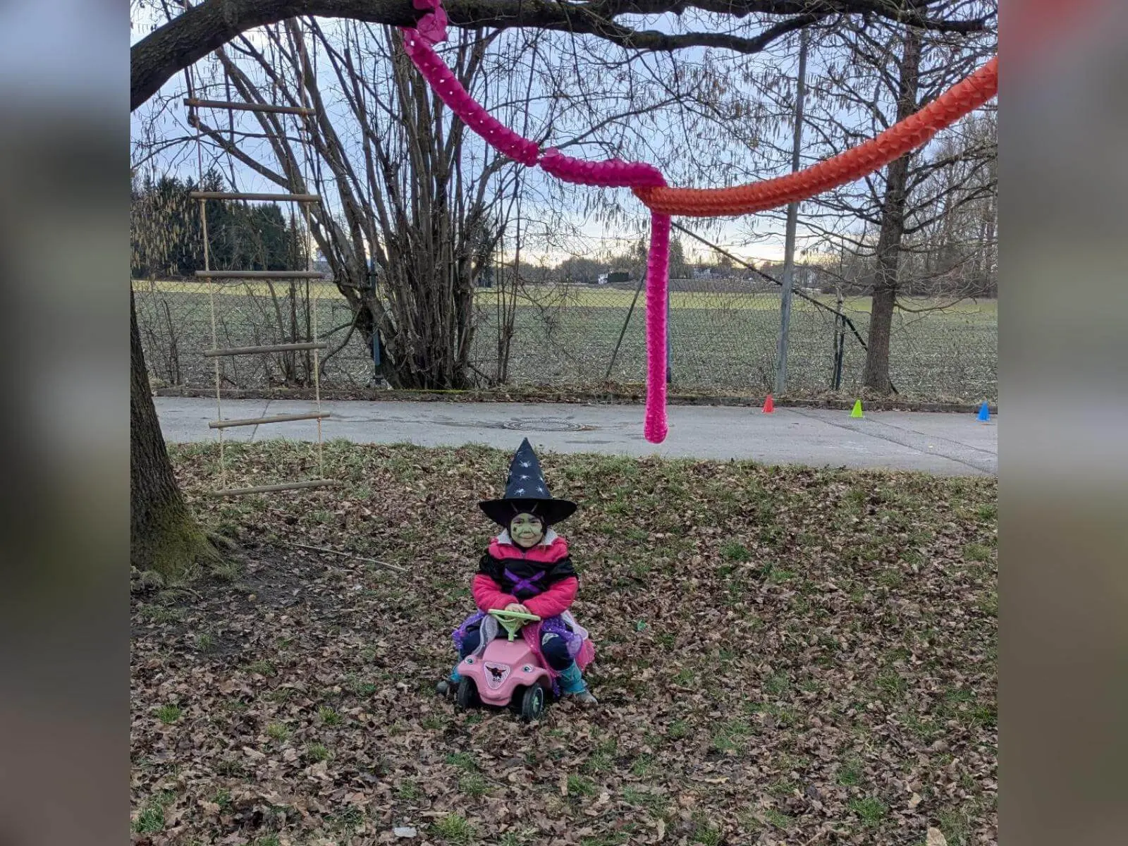 Ein verkleidetes kleines Kind mit Hexenhut sitzt auf einem Rutschfahrzeug unter bunten Girlanden an einem Baum auf einer Wiese. | © DAV Markt Schwaben
