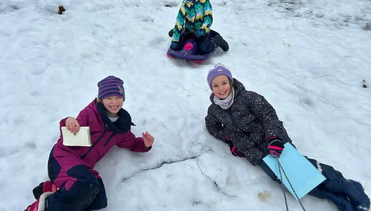 Drei Kinder spielen im Schnee; zwei liegen lachend vorn mit Notizbuch und Mappe, hinten sitzt ein Kind auf einem Schlitten am Fundort ihrer Entdeckung. | © DAV Markt Schwaben, Familiengruppe