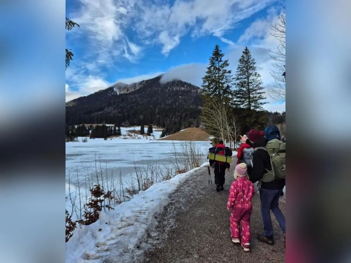 Kinder und Erwachsene gehen am verschneiten Uferweg entlang eines gefrorenen Sees; im Hintergrund erheben sich Berge und Wald unter blauem Himmel. | © DAV Markt Schwaben, Familiengruppe