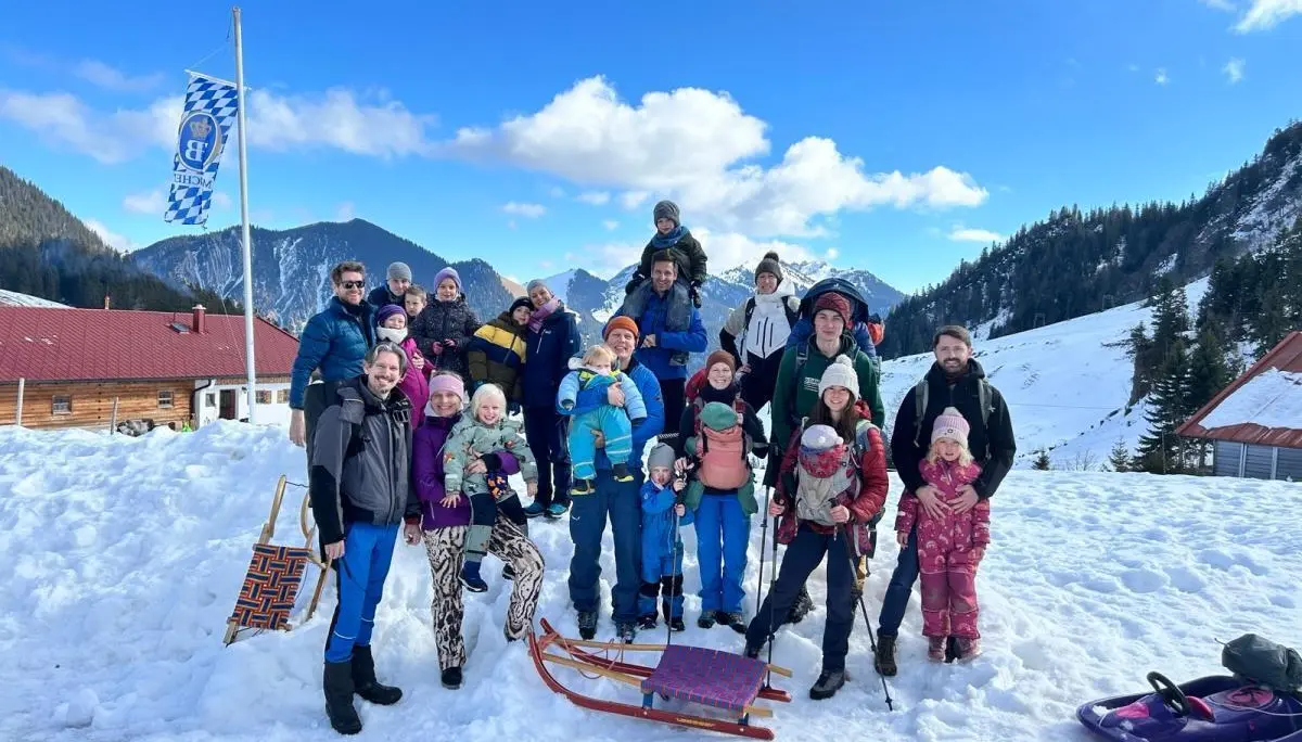 Kinder und Erwachsene stehen und sitzen im Schnee vor Bergkulisse; Schlitten liegen davor, alle tragen Winterkleidung unter blauem Himmel. | © DAV Markt Schwaben