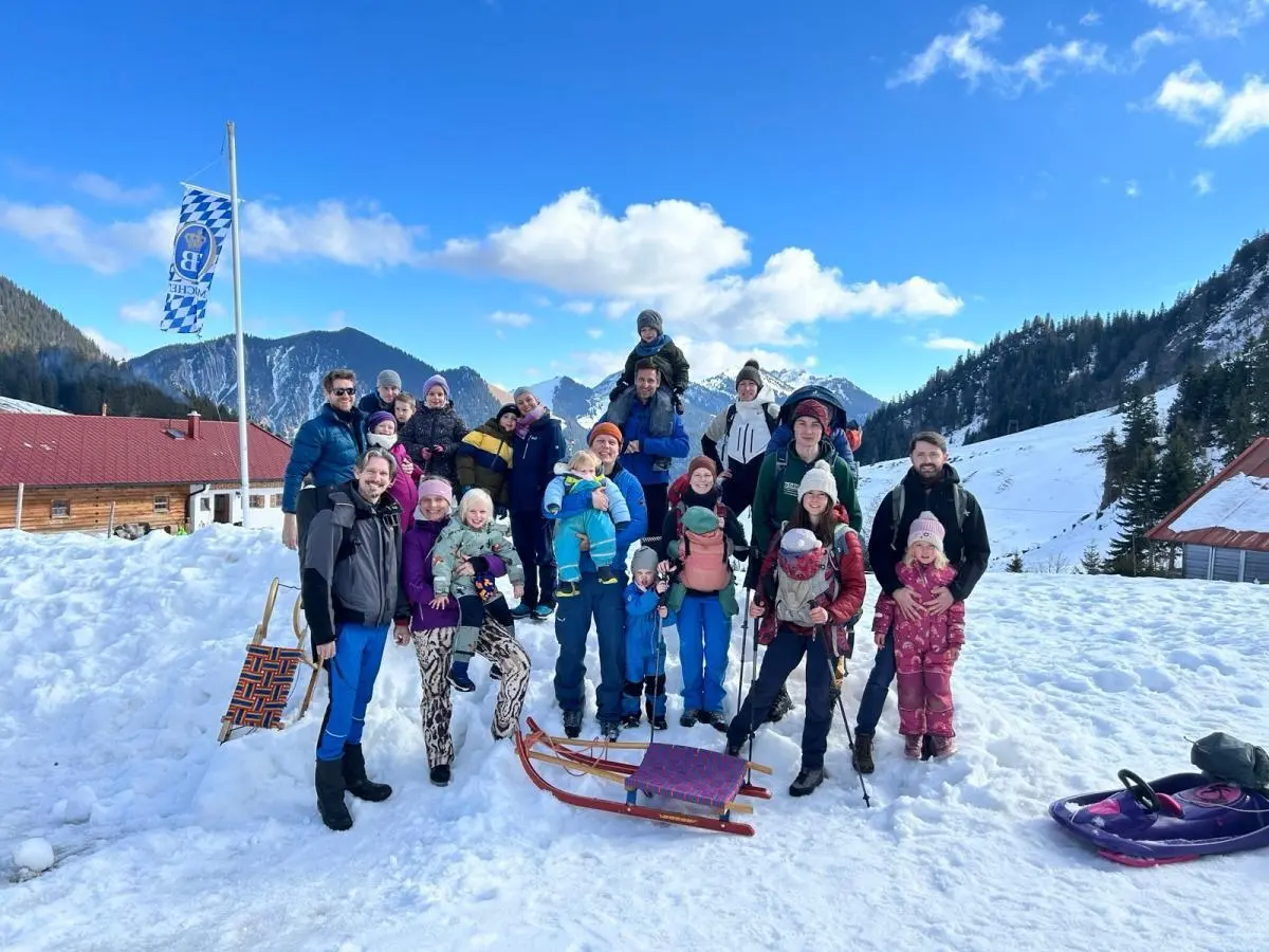 Kinder und Erwachsene stehen und sitzen im Schnee vor Bergkulisse; Schlitten liegen davor, alle tragen Winterkleidung unter blauem Himmel. | © DAV Markt Schwaben