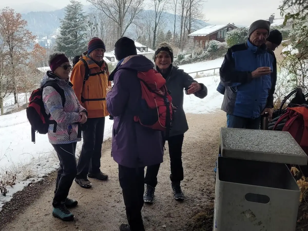 Sechs Wandernde stehen auf einem verschneiten Weg; vorne ein Abfallbehälter, dahinter Häuser, Bäume und winterliche Hänge.  | © DAV Markt Schwaben / Hubert Inhofer