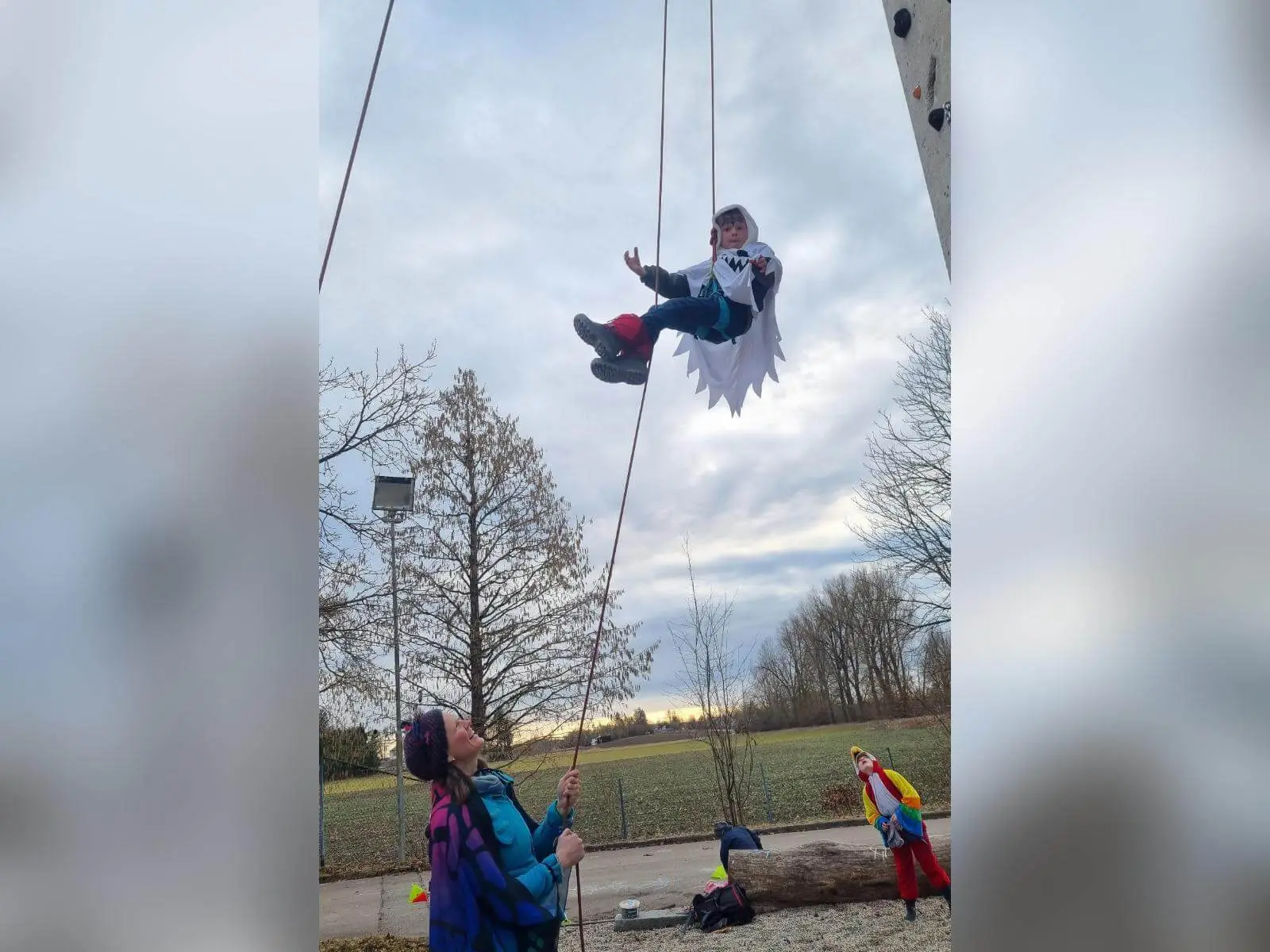 Ein verkleidetes Kind hängt gesichert an einem Kletterseil in der Luft, unten sichern Erwachsene und Kinder in bunten Kostümen. | © DAV Markt Schwaben