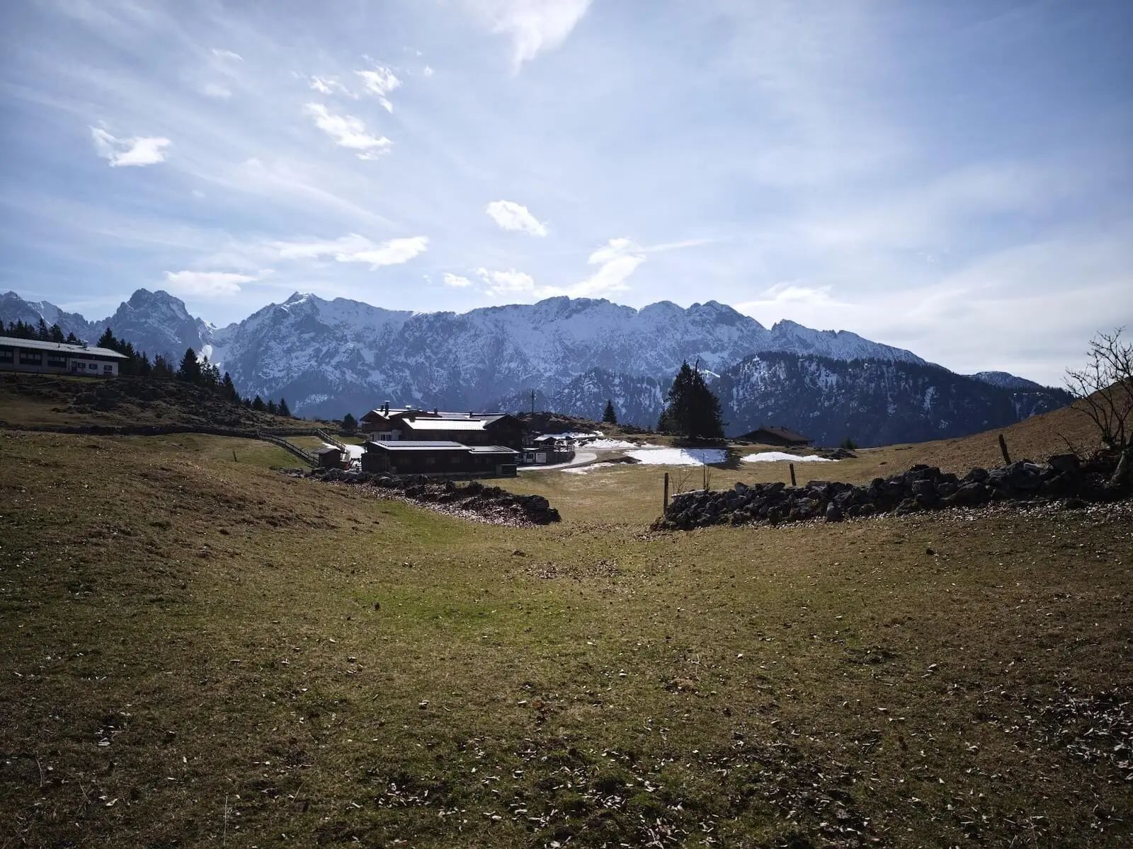 Grüne Wiesen mit Schneeflecken, eine Steinmauer und mehrere Almhäuser liegen vor schneebedeckten Bergen unter klarem Himmel. | © DAV Markt Schwaben/Erwin Matzinger