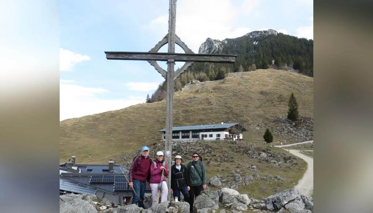Vier Wandernde stehen auf felsigem Gelände neben einem großen Holzkreuz, dahinter liegen Almgebäude und bewaldete Berghänge. | © DAV Markt Schwaben/Gerlinde Hübl