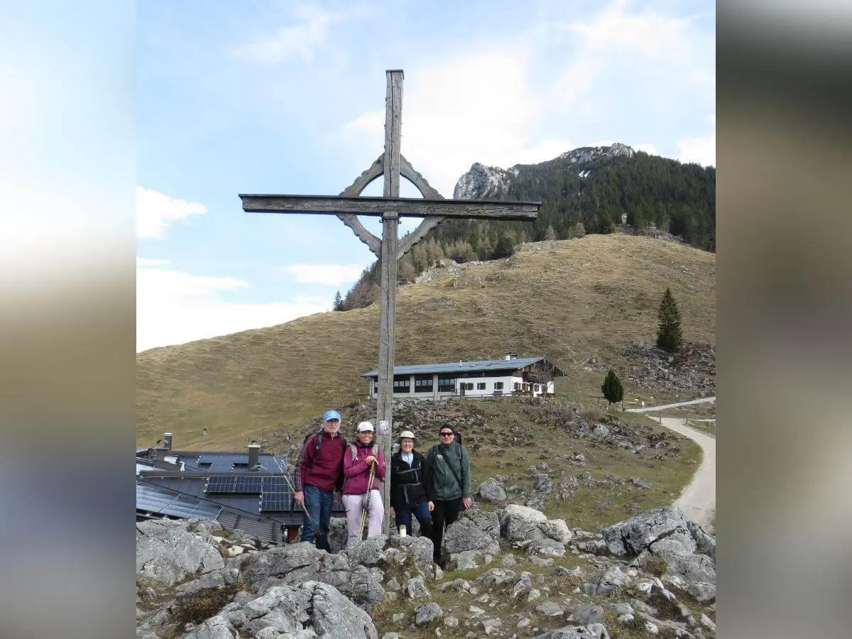 Vier Wandernde stehen auf felsigem Gelände neben einem großen Holzkreuz, dahinter liegen Almgebäude und bewaldete Berghänge. | © DAV Markt Schwaben/Gerlinde Hübl