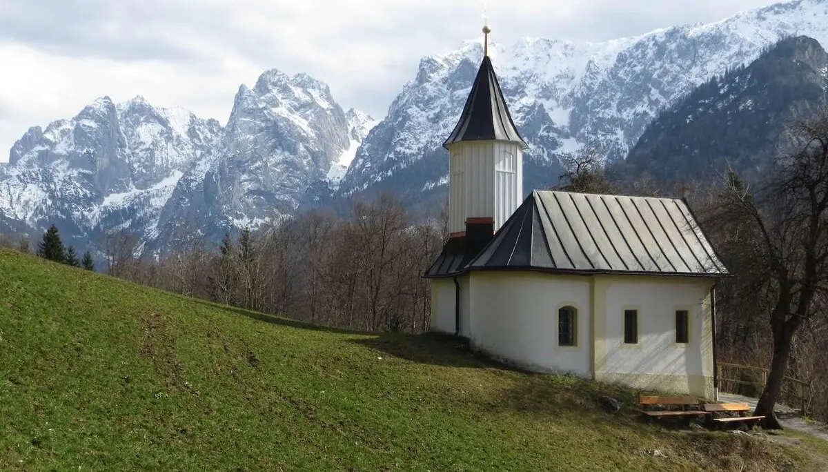 Eine kleine weiße Kapelle mit Turm steht auf einer grünen Anhöhe, dahinter erheben sich schneebedeckte Berge unter blauem Himmel. | © DAV Markt Schwaben/Gerlinde Hübl