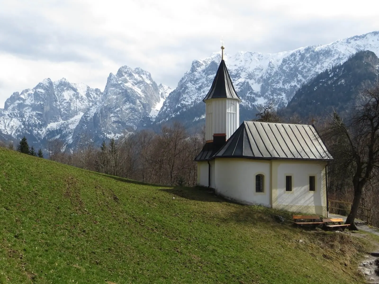 Eine kleine weiße Kapelle mit Turm steht auf einer grünen Anhöhe, dahinter erheben sich schneebedeckte Berge unter blauem Himmel. | © DAV Markt Schwaben/Gerlinde Hübl
