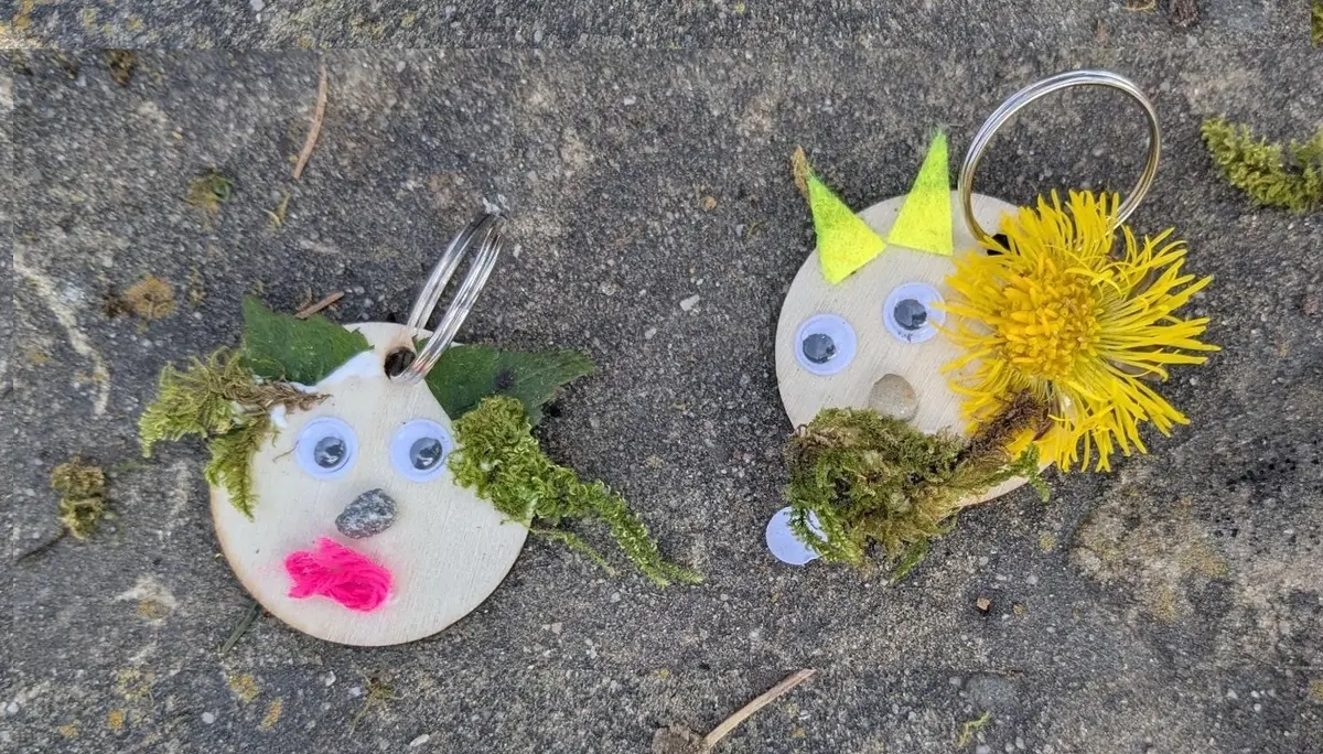 Zwei runde Holzanhänger liegen auf grauem Untergrund und zeigen mit Naturmaterialien gestaltete Trollgesichter mit Moos, Augen und Blüten. | © DAV Markt Schwaben/Silke Titze