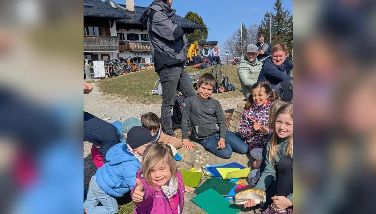 Kinder sitzen draußen vor einer Hütte auf dem Boden und basteln mit Papier und Materialien, während Erwachsene im Hintergrund zuschauen. | © DAV Markt Schwaben/Silke Titze