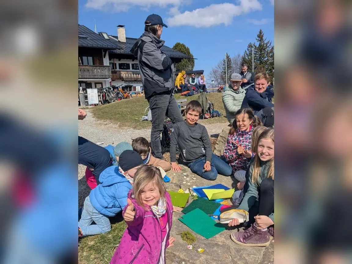 Kinder sitzen draußen vor einer Hütte auf dem Boden und basteln mit Papier und Materialien, während Erwachsene im Hintergrund zuschauen. | © DAV Markt Schwaben/Silke Titze