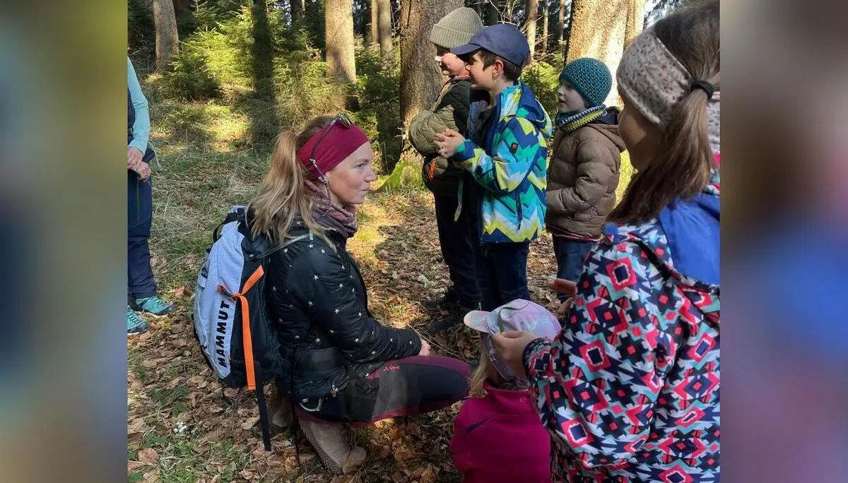 Ein Erwachsener kniet im Wald vor mehreren Kindern und erklärt ihnen etwas, während alle warm gekleidet zwischen hohen Bäumen stehen. | © DAV Markt Schwaben/Silke Titze