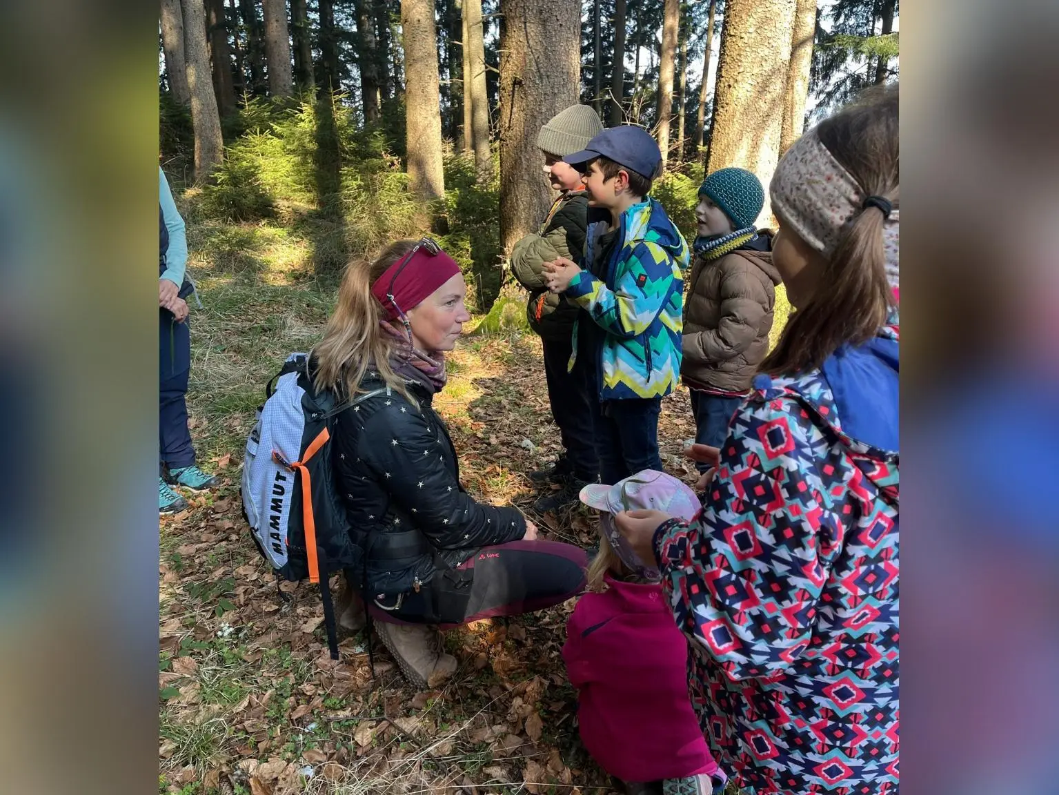 Ein Erwachsener kniet im Wald vor mehreren Kindern und erklärt ihnen etwas, während alle warm gekleidet zwischen hohen Bäumen stehen. | © DAV Markt Schwaben/Silke Titze