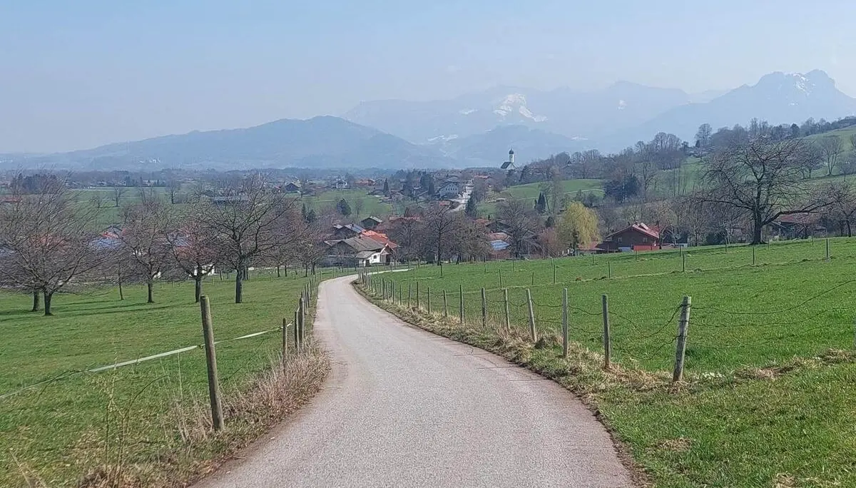Schmaler asphaltierter Weg führt durch grüne Wiesen und Streuobstflächen auf einen Ort zu, im Hintergrund Hügel und Berge bei klarer Sicht. | © DAV Markt Schwaben