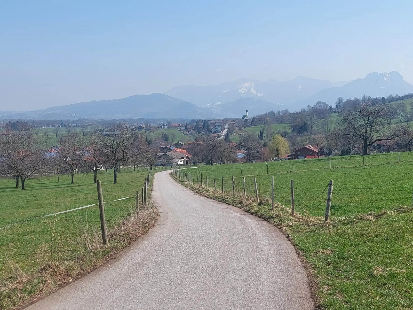 Schmaler asphaltierter Weg führt durch grüne Wiesen und Streuobstflächen auf einen Ort zu, im Hintergrund Hügel und Berge bei klarer Sicht. | © DAV Markt Schwaben