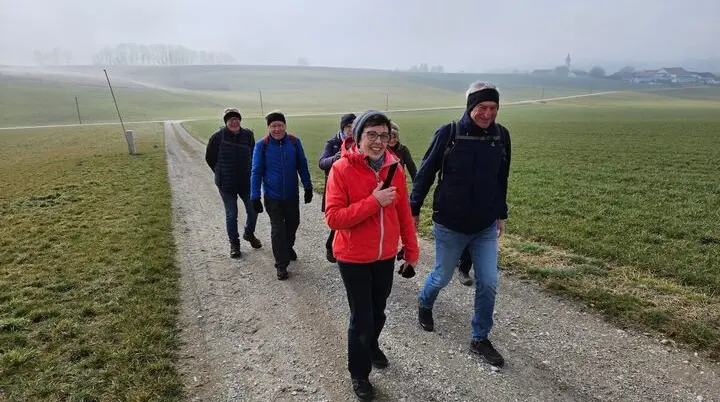 Sechs Wandernde gehen auf einem Kiesweg durch offene Felder; vorne die Gruppe, dahinter Hügel, Bäume und ein Dorf mit Kirchturm im Dunst.   | © DAV Markt Schwaben/Hubert Inhofer