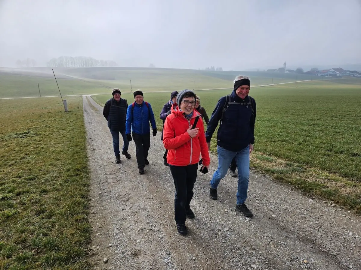 Sechs Wandernde gehen auf einem Kiesweg durch offene Felder; vorne die Gruppe, dahinter Hügel, Bäume und ein Dorf mit Kirchturm im Dunst.   | © DAV Markt Schwaben/Hubert Inhofer