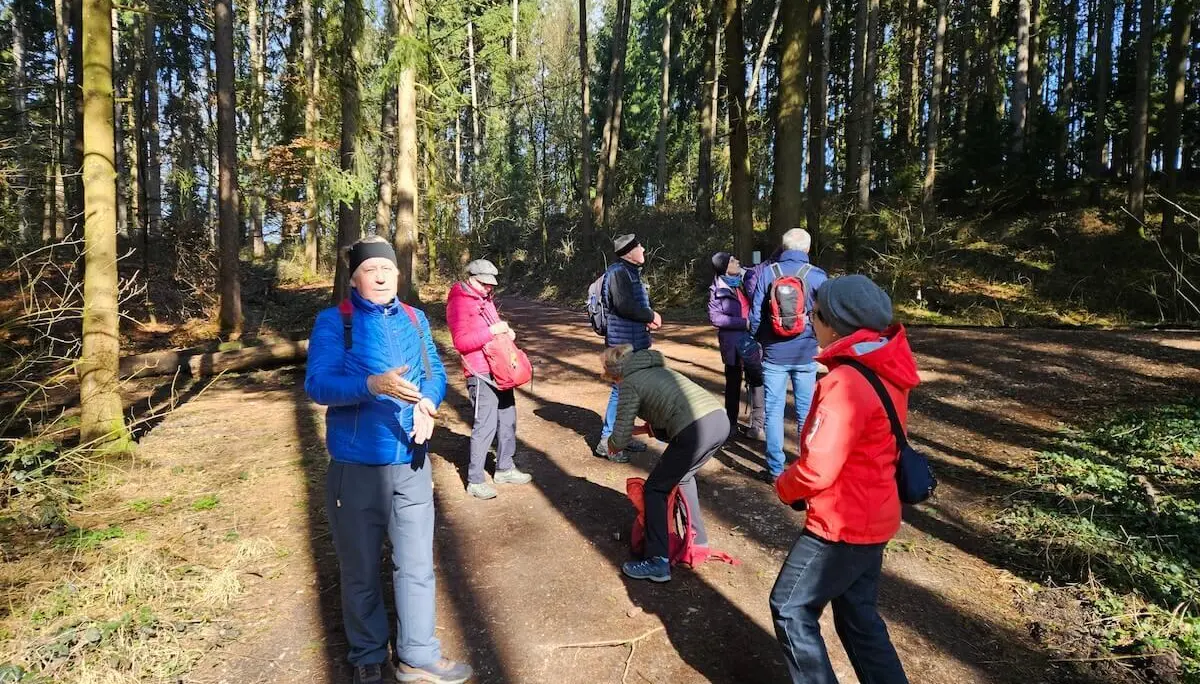 Mehrere Wandernde stehen auf einem Waldweg; vorne Personen im Gespräch, dahinter hohe Bäume und Sonnenlicht zwischen den Stämmen. | © DAV Markt Schwaben/Hubert Inhofer
