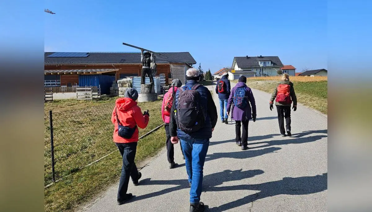 Sieben Wandernde gehen auf einer Landstraße an Häusern vorbei; vorne die Gruppe, rechts eine Statue auf Sockel, oben ein kleines Flugzeug am Himmel. | © DAV Markt Schwaben/Hubert Inhofer
