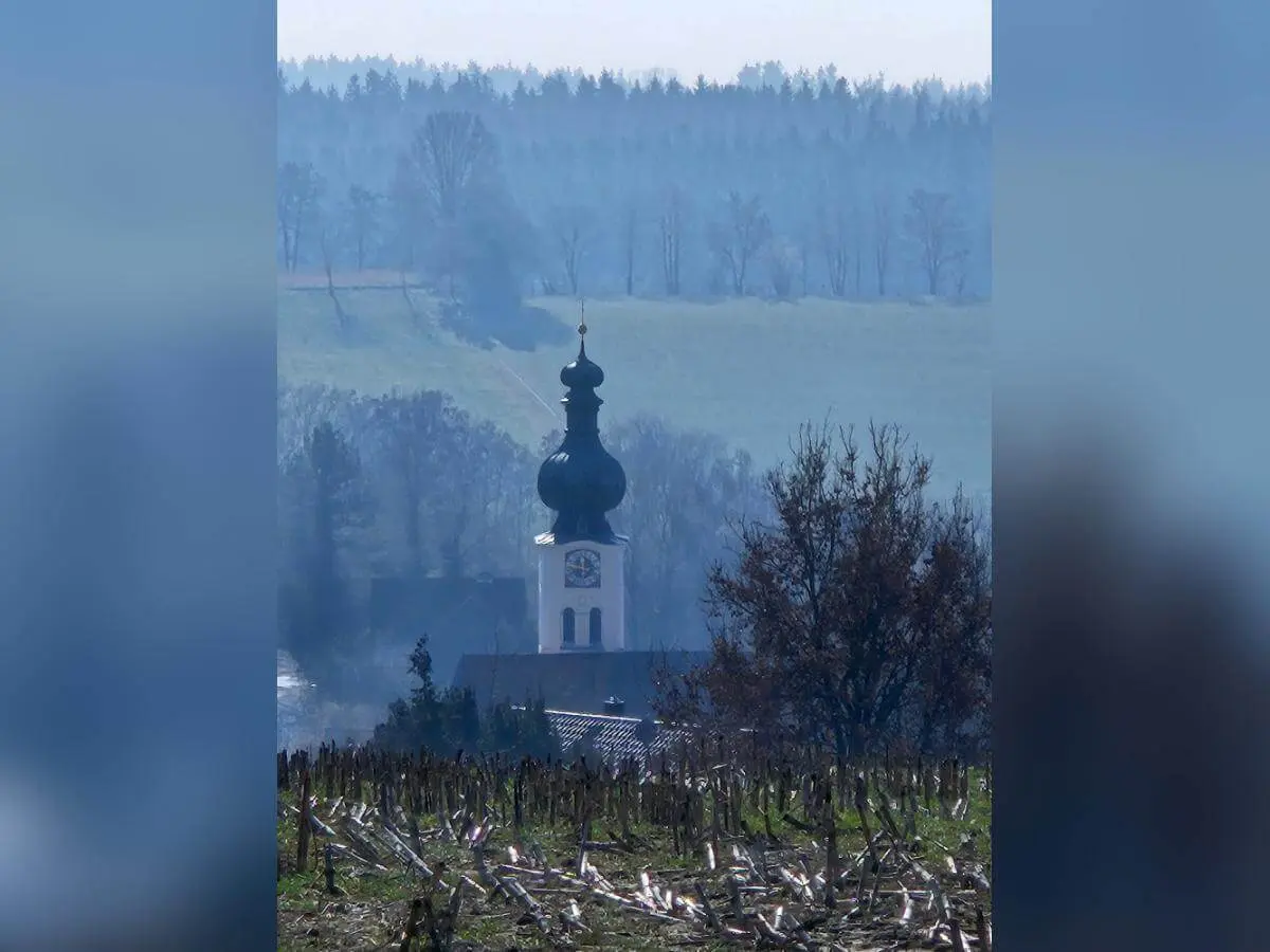 Eine Kirche mit Zwiebelturm steht in einem Tal; vorne ein abgeerntetes Feld, dahinter Hügel und Wald, teils im dunstigen Licht. | © DAV Markt Schwaben/Hubert Inhofer