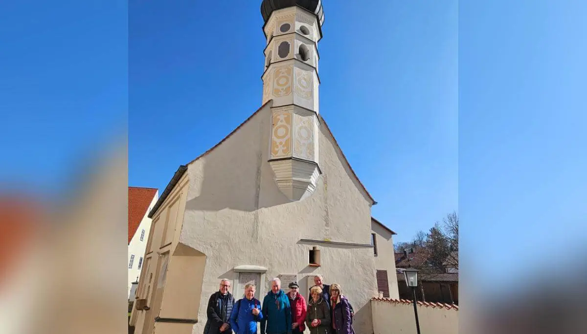Sechs Wandernde stehen vor einer kleinen Kirche mit Turm; vorne die Gruppe, dahinter helle Fassaden und klarer Himmel.  | © DAV Markt Schwaben/Hubert Inhofer