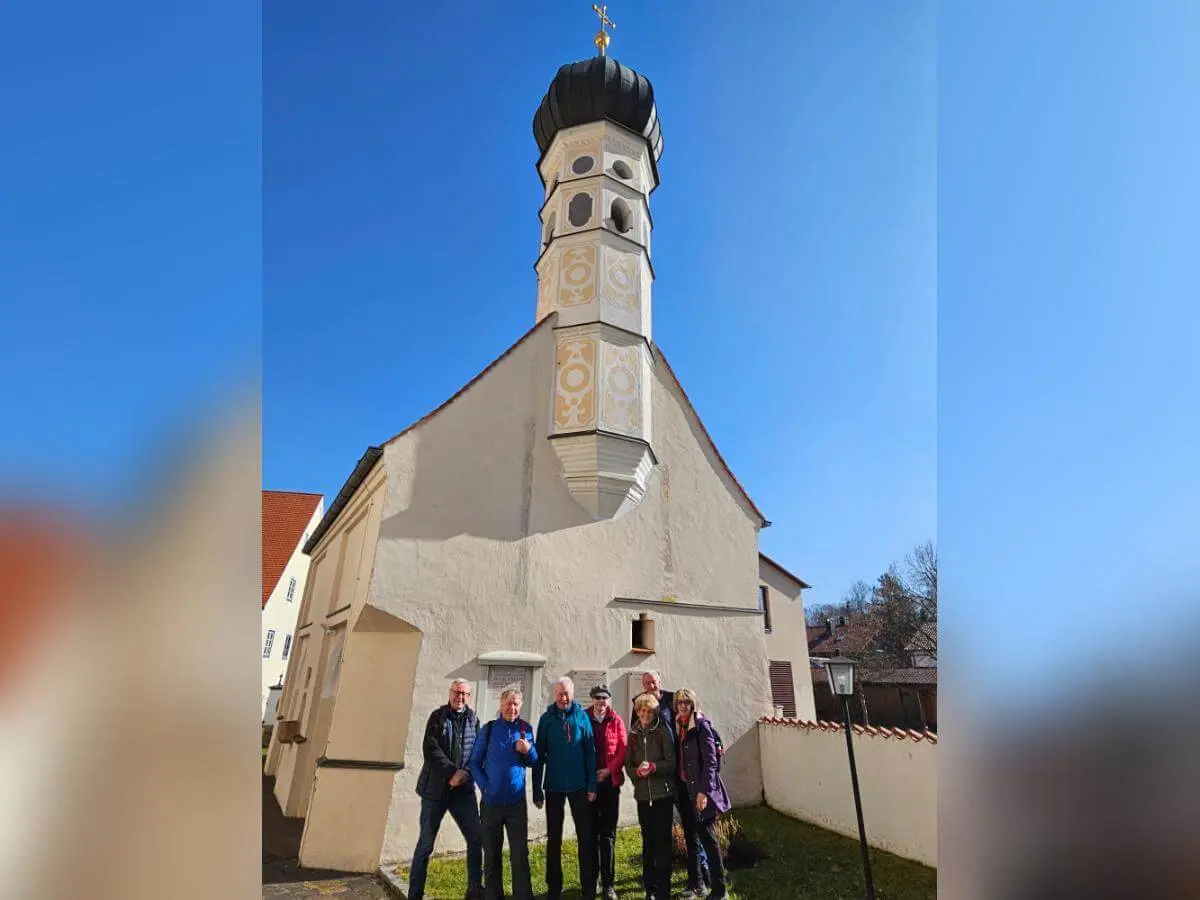 Sechs Wandernde stehen vor einer kleinen Kirche mit Turm; vorne die Gruppe, dahinter helle Fassaden und klarer Himmel.  | © DAV Markt Schwaben/Hubert Inhofer