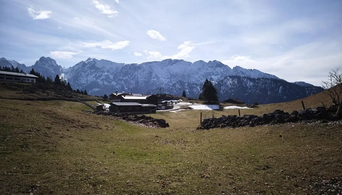 Grüne Wiesen mit Schneeflecken, eine Steinmauer und mehrere Almhäuser liegen vor schneebedeckten Bergen unter klarem Himmel. | © DAV Markt Schwaben/Erwin Matzinger