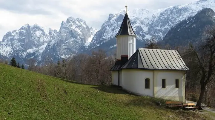 Eine kleine weiße Kapelle mit Turm steht auf einer grünen Anhöhe, dahinter erheben sich schneebedeckte Berge unter blauem Himmel. | © DAV Markt Schwaben/Gerlinde Hübl