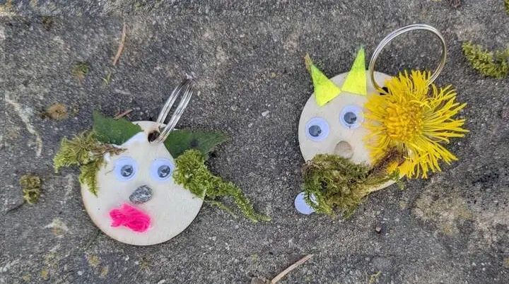 Zwei runde Holzanhänger liegen auf grauem Untergrund und zeigen mit Naturmaterialien gestaltete Trollgesichter mit Moos, Augen und Blüten. | © DAV Markt Schwaben/Silke Titze