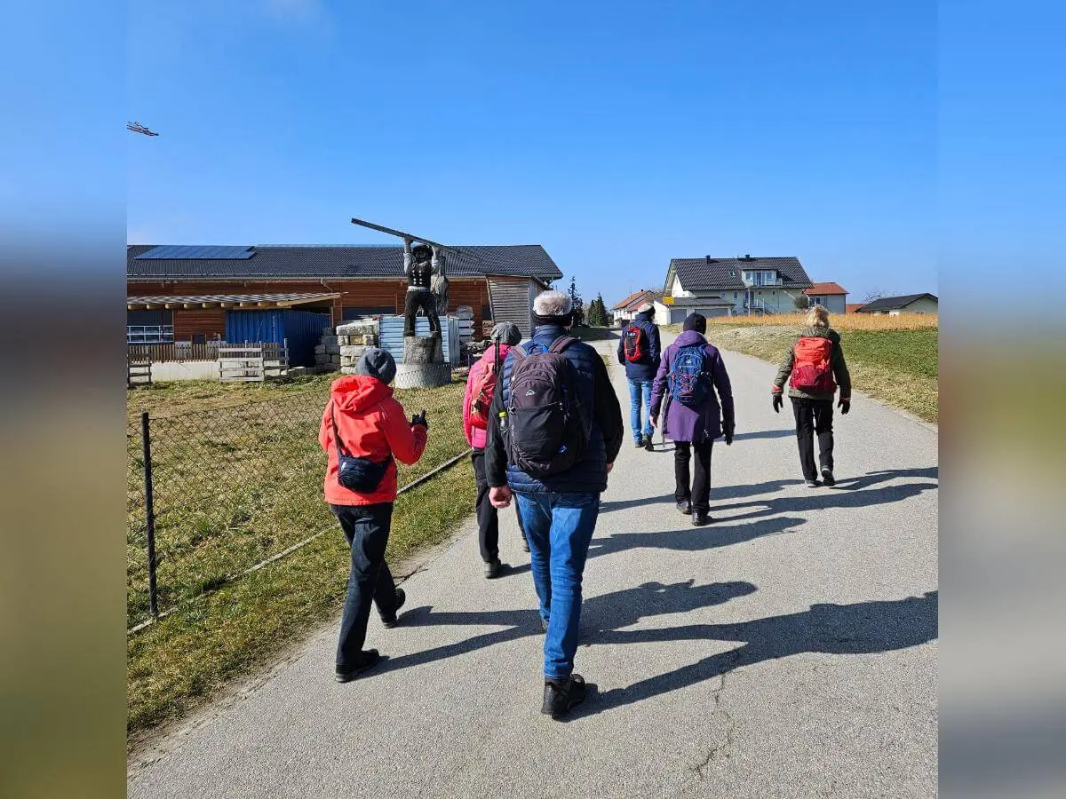 Sieben Wandernde gehen auf einer Landstraße an Häusern vorbei; vorne die Gruppe, rechts eine Statue auf Sockel, oben ein kleines Flugzeug am Himmel. | © DAV Markt Schwaben / Hubert Inhofer