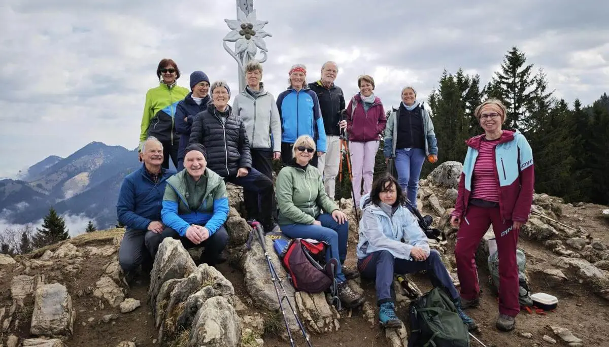 Eine Wandergruppe sitzt und steht auf felsigem Gipfelgelände vor einem Gipfelkreuz, umgeben von Bergen und Wald. | © DAV Markt Schwaben/Erwin Matzinger