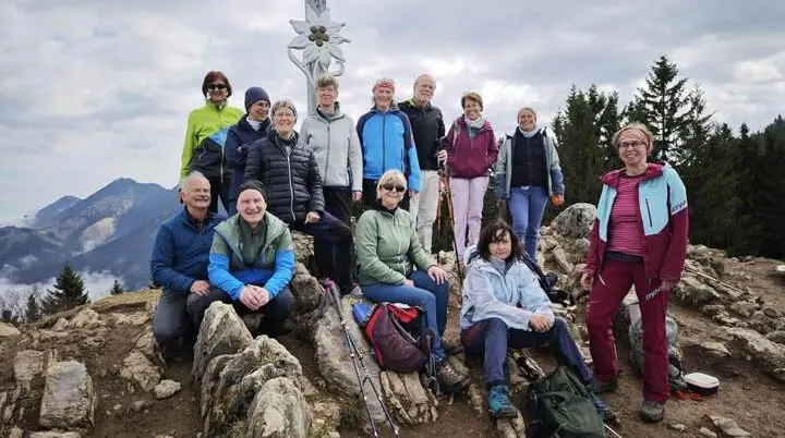 Eine Wandergruppe sitzt und steht auf felsigem Gipfelgelände vor einem Gipfelkreuz, umgeben von Bergen und Wald. | © DAV Markt Schwaben/Erwin Matzinger