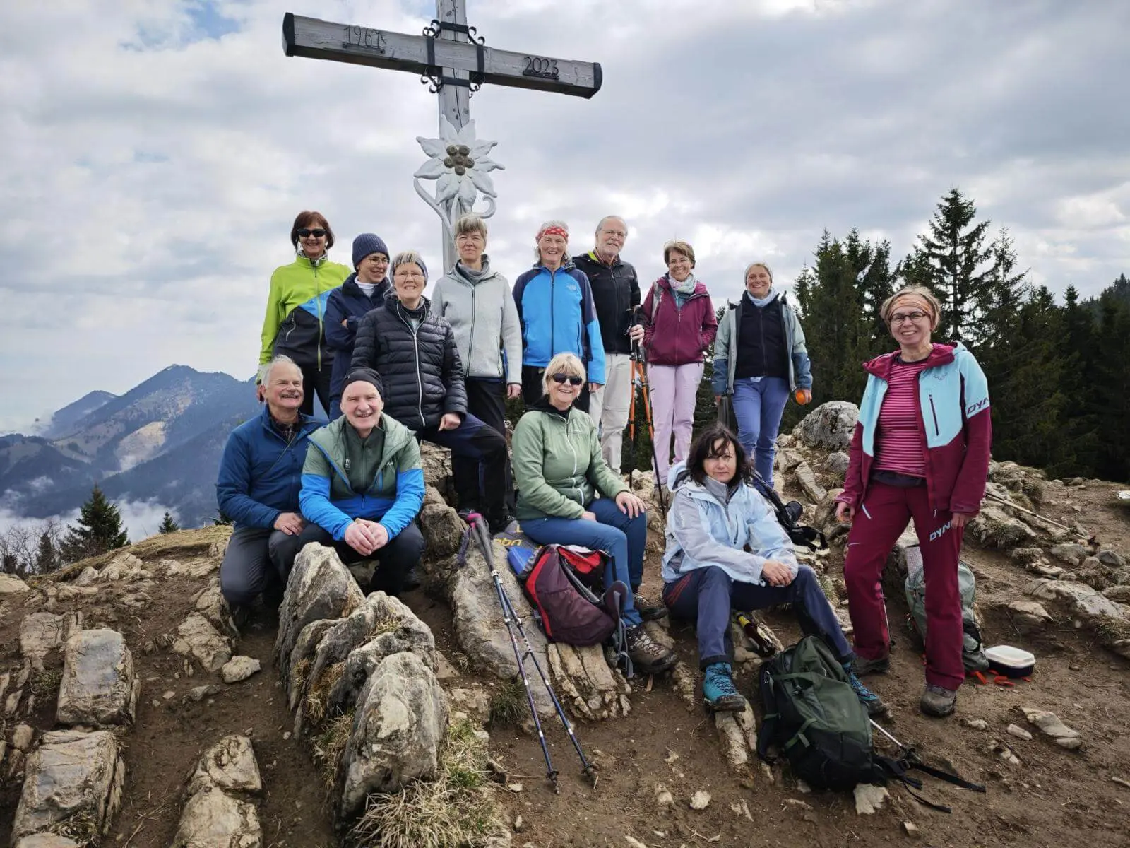 Eine Wandergruppe sitzt und steht auf felsigem Gipfelgelände vor einem Gipfelkreuz, umgeben von Bergen und Wald. | © DAV Markt Schwaben/Erwin Matzinger