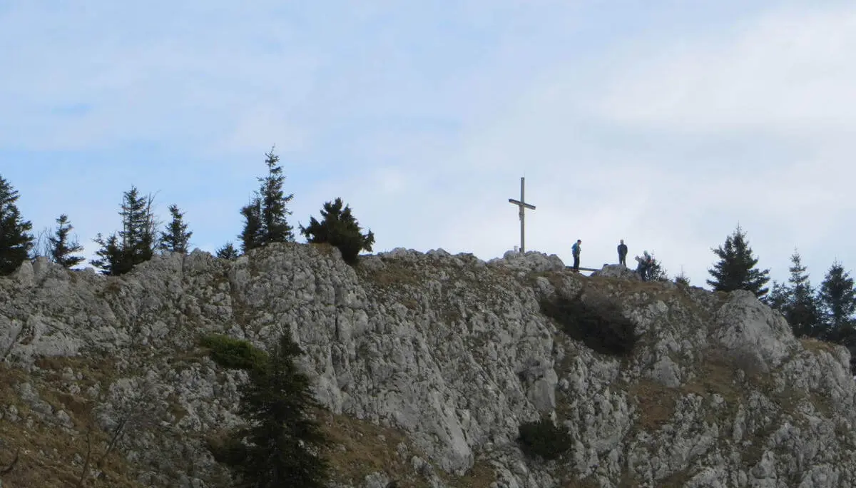 Auf einem felsigen Grat steht ein Gipfelkreuz; daneben pausieren mehrere Wandernde, umgeben von Felsen, Bäumen und Himmel. | © DAV Markt Schwaben/Gerlinde Hübl