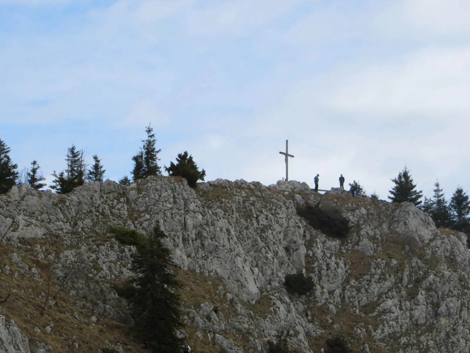 Auf einem felsigen Grat steht ein Gipfelkreuz; daneben pausieren mehrere Wandernde, umgeben von Felsen, Bäumen und Himmel. | © DAV Markt Schwaben/Gerlinde Hübl