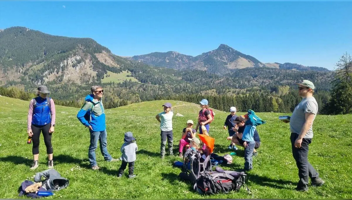 Familiengruppe mit Kindern macht Pause auf grüner Almwiese; Rucksäcke liegen am Boden, im Hintergrund Bergpanorama. | © DAV Markt Schwaben/Maria Wenisch